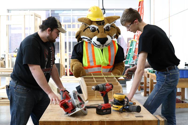 Willy the Wildcat works with students in a woodworking lab