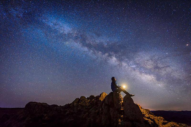 A person sits on a rock formation and looks up at the Milky Way
