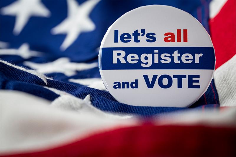 A photo of a button saying "Let's all register and vote" sitting on an American flag.